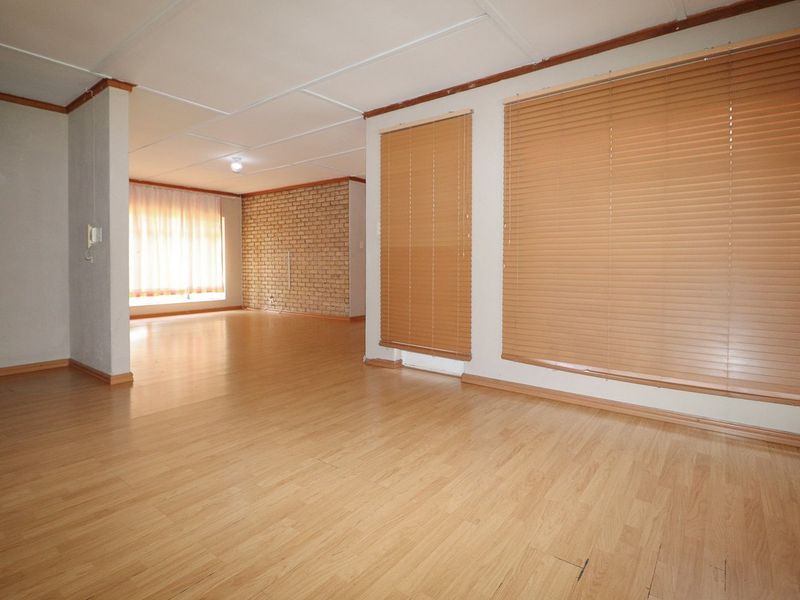 Spacious, empty living area featuring laminate flooring, an exposed brick feature wall, and large windows with fitted venetian blinds.