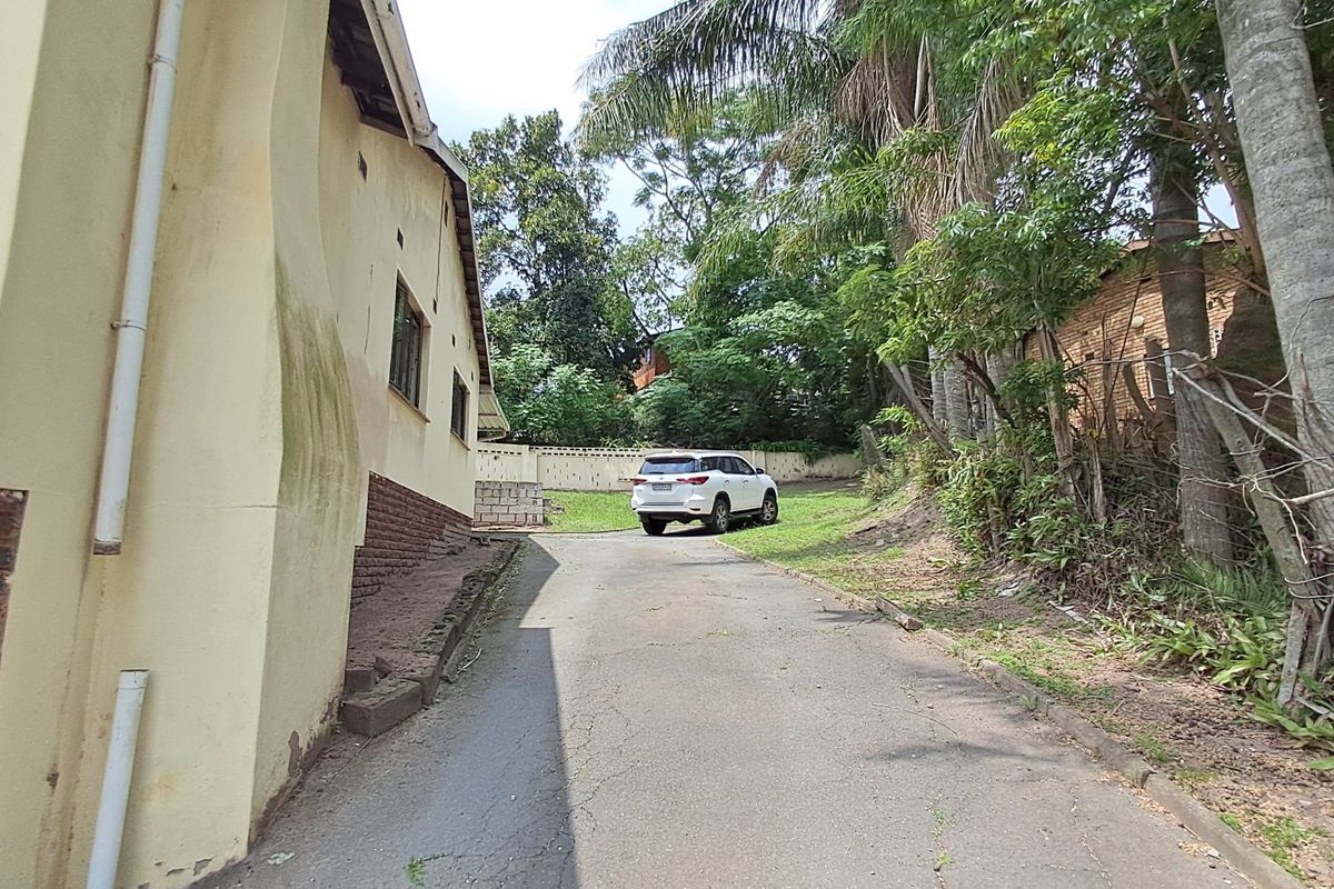 A view of the rear exterior area featuring an asphalt driveway, brick retaining wall, and mature trees surrounding the property.
