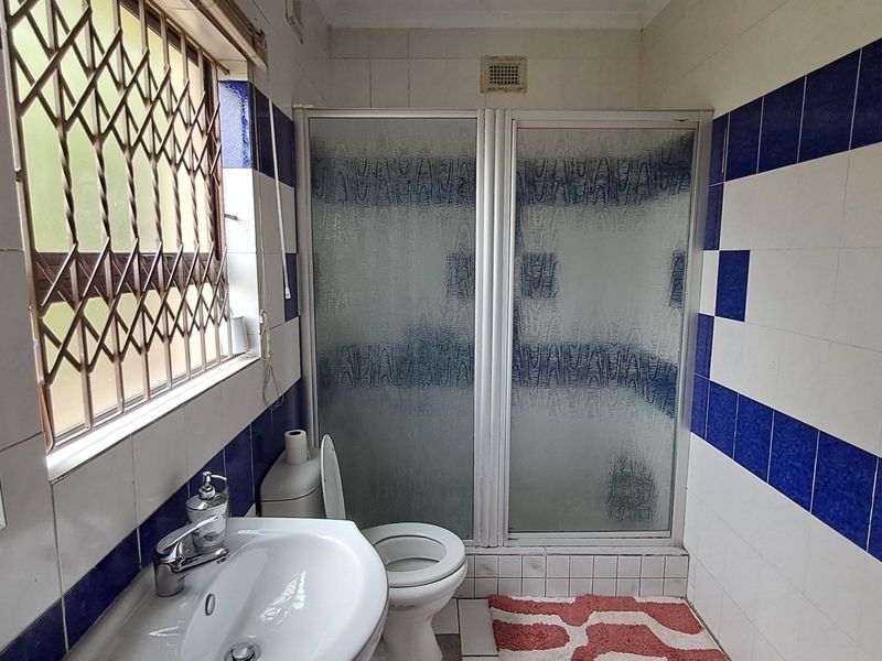 A functional bathroom featuring a shower enclosure, pedestal basin, and toilet, finished with dated blue and white wall tiles and a security window gate.
