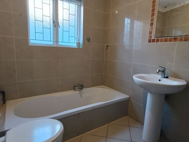 A functional bathroom featuring a bathtub, pedestal basin, and neutral wall tiling with a window providing natural light.