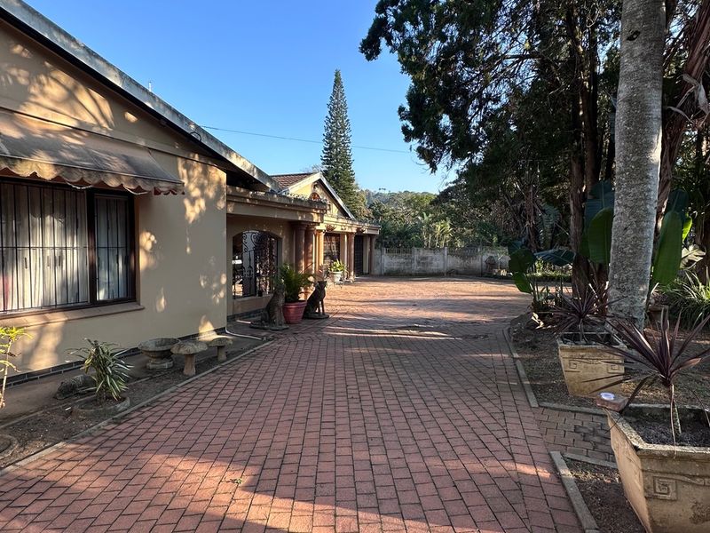 A view of the front exterior featuring a brick paved driveway leading towards the house entrance, complemented by mature trees and a perimeter wall.