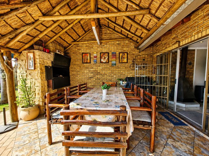 Covered outdoor patio featuring a traditional thatched roof, exposed brickwork, stone-clad built-in braai, and tiled flooring, providing a rustic space for outdoor dining.