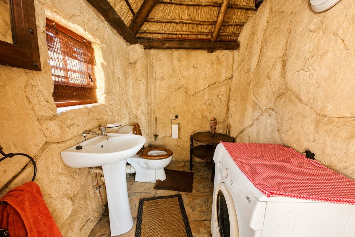 A rustic bathroom featuring stone-textured walls, a thatched roof, pedestal basin, and toilet, with integrated laundry space.