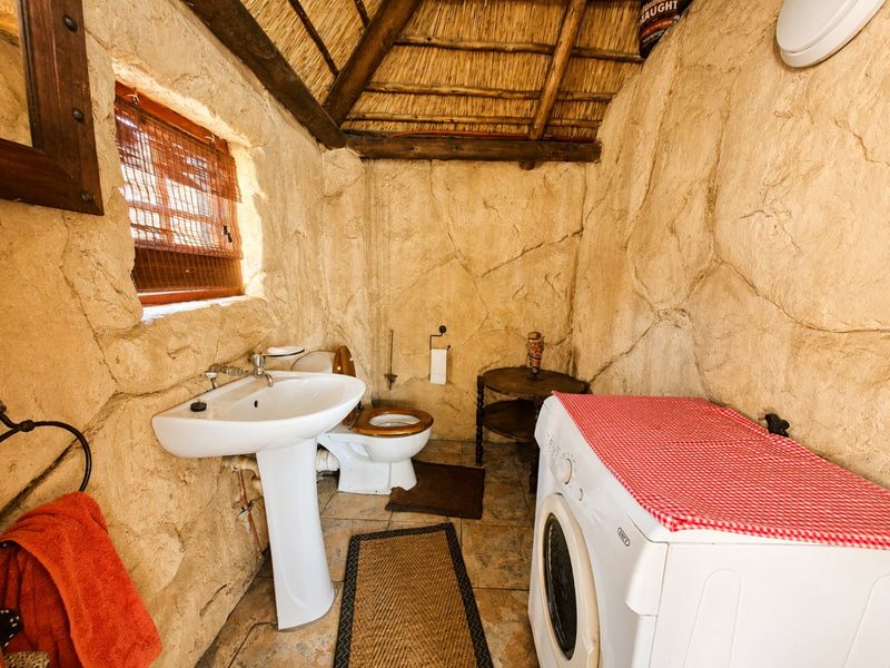 A rustic bathroom featuring stone-textured walls, a thatched roof, pedestal basin, and toilet, with integrated laundry space.