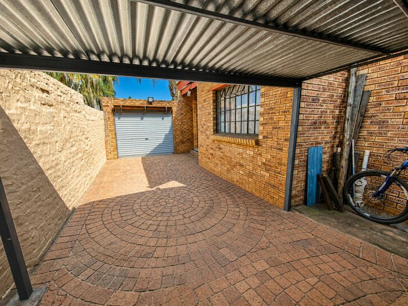 A paved driveway area featuring a corrugated metal carport structure, brick boundary walls, and a single roll-up garage door.