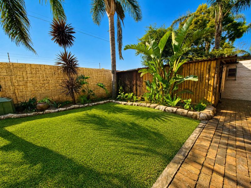 A well-maintained rear garden featuring an artificial lawn, brick paved walkway, and a combination of stone-clad and timber boundary fencing.