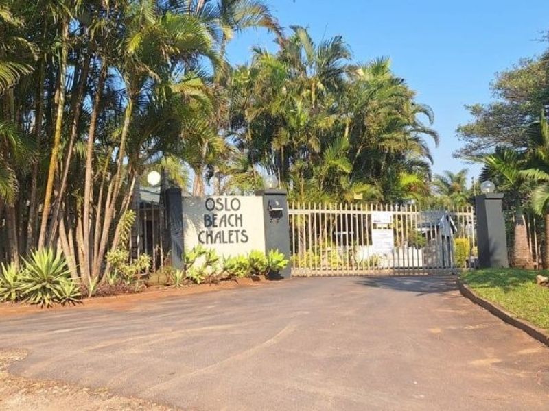 The entrance to Oslo Beach Chalets featuring a secure gated access point, paved driveway, and established tropical landscaping.