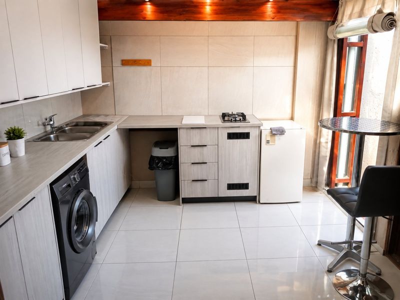 Compact modern kitchen featuring light wood-grain cabinetry, stainless steel sink, gas hob, tiled flooring, and an exposed timber beam ceiling detail.