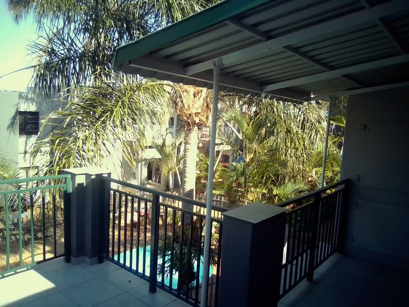 A covered balcony featuring tiled flooring and metal balustrades, overlooking the complex garden and swimming pool area.