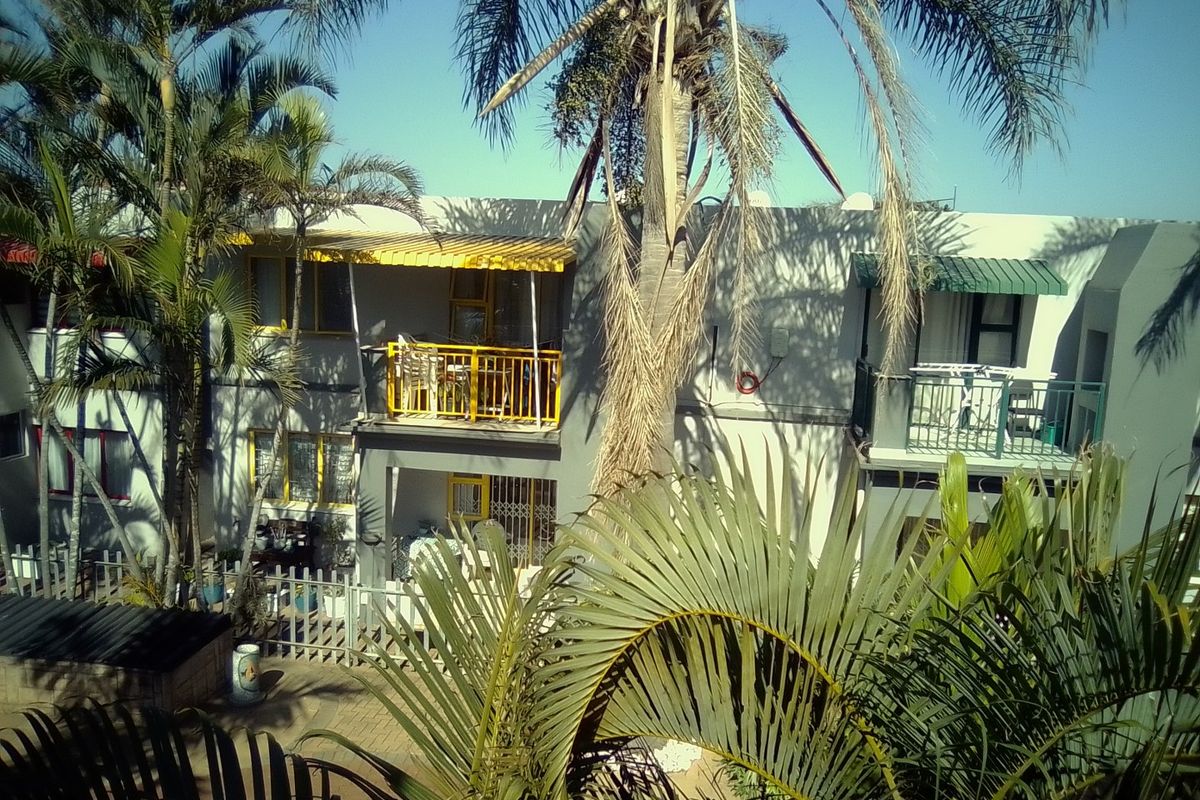 Rear view of an apartment complex featuring balconies with metal railings, awnings, and a paved courtyard area surrounded by palm trees.