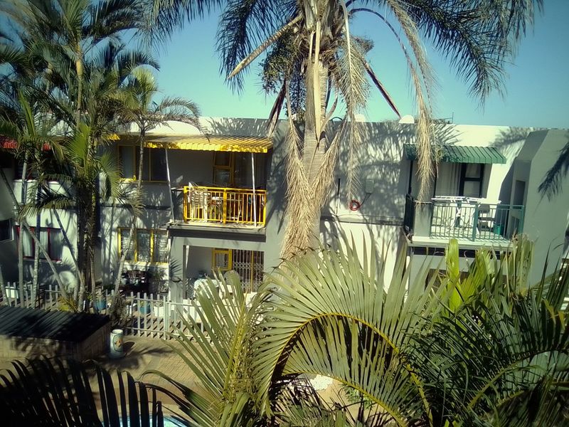 Rear view of an apartment complex featuring balconies with metal railings, awnings, and a paved courtyard area surrounded by palm trees.
