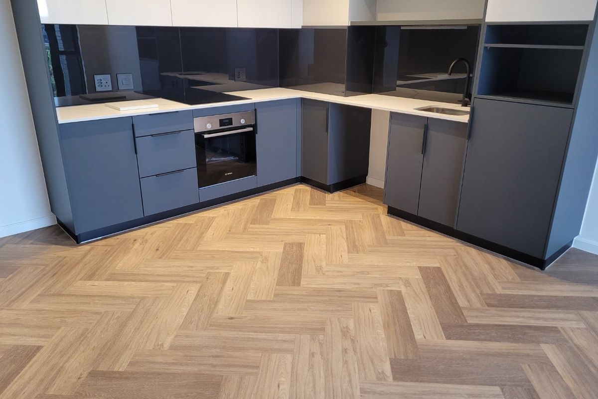 Modern kitchen featuring sleek grey and white cabinetry, integrated oven, electric hob, glass splashback, and stylish herringbone wood-look flooring.