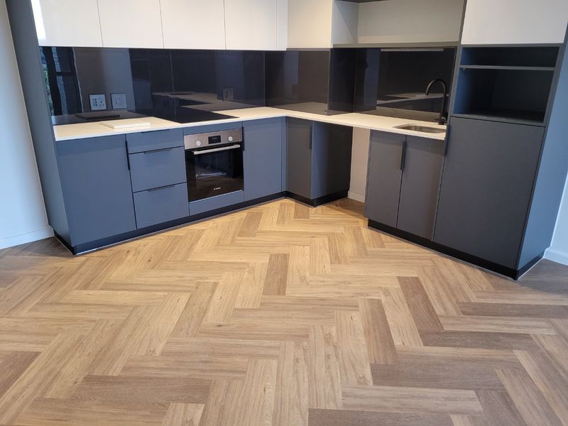 Modern kitchen featuring sleek grey and white cabinetry, integrated oven, electric hob, glass splashback, and stylish herringbone wood-look flooring.