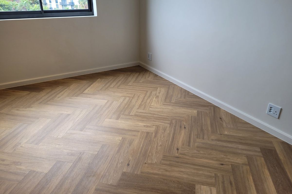 A bright, empty bedroom featuring modern herringbone timber flooring, clean white walls, and a large window with an aluminium frame.