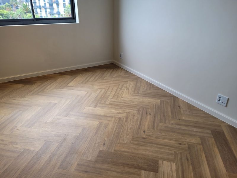 A bright, empty bedroom featuring modern herringbone timber flooring, clean white walls, and a large window with an aluminium frame.