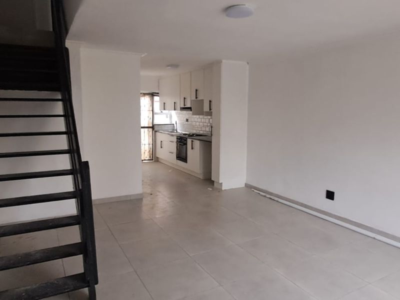 Open plan living area featuring a staircase and a compact kitchen with built-in cabinetry, oven, and hob, finished with neutral floor tiles.