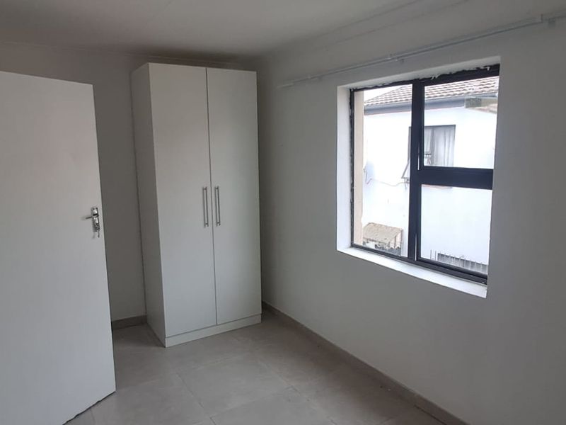 A simple, neutral bedroom featuring tiled flooring, a built-in wardrobe, and a window providing natural light.