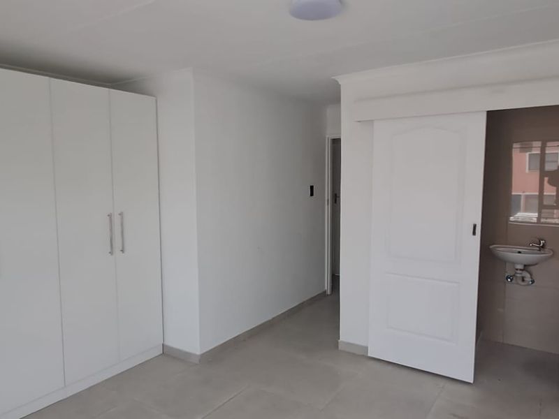 A clean, modern bedroom featuring built-in cupboards, neutral tiled flooring, and a sliding door leading to an en-suite bathroom.