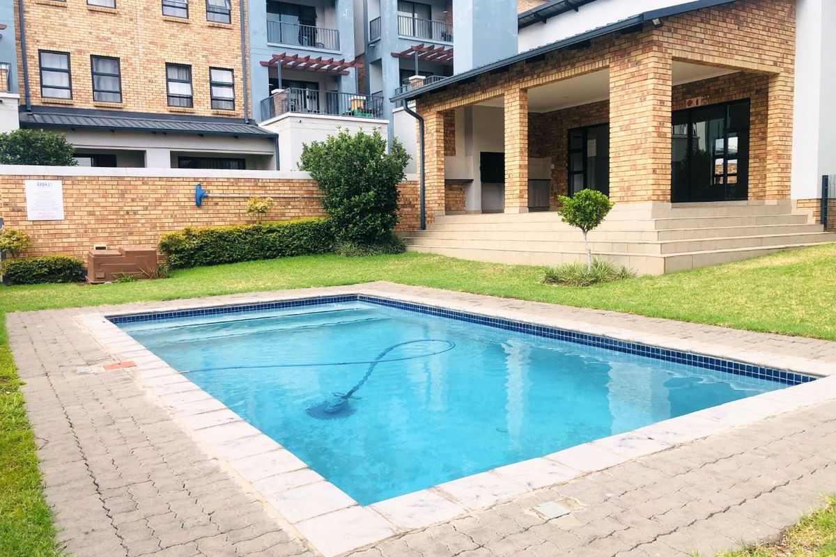A well-maintained communal pool area featuring a paved surround, manicured lawn, and a covered patio area within a brick-walled apartment complex.