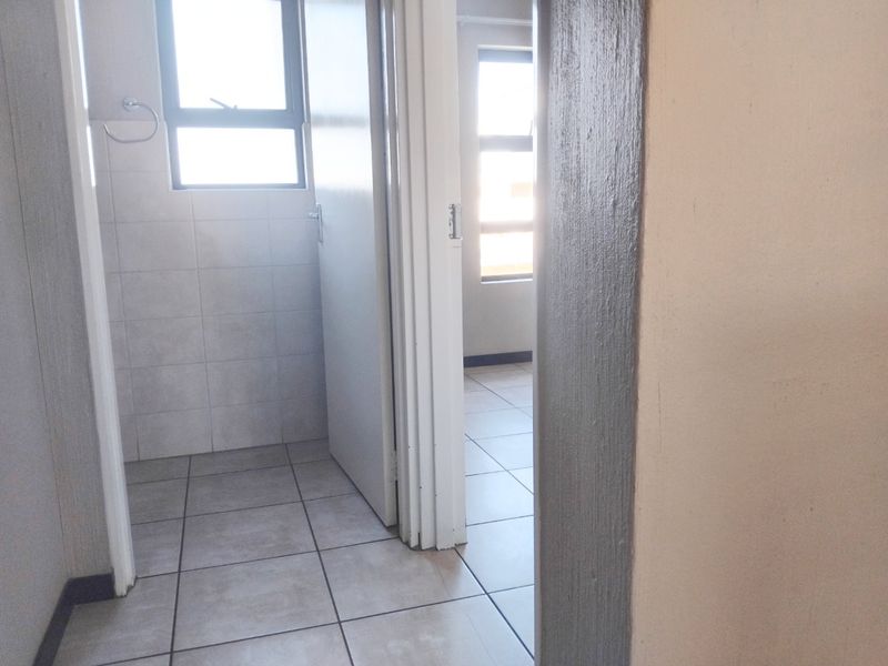 A view through a hallway connecting to a bathroom and another room, featuring neutral tiled flooring and aluminium window frames.