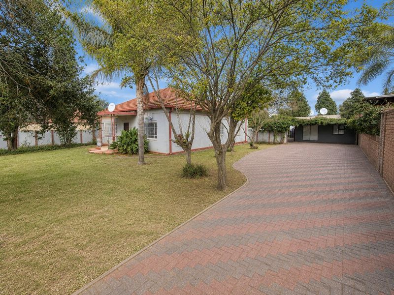 Front view of a single-story house featuring a large lawn, extensive paved driveway, and secure boundary walling.
