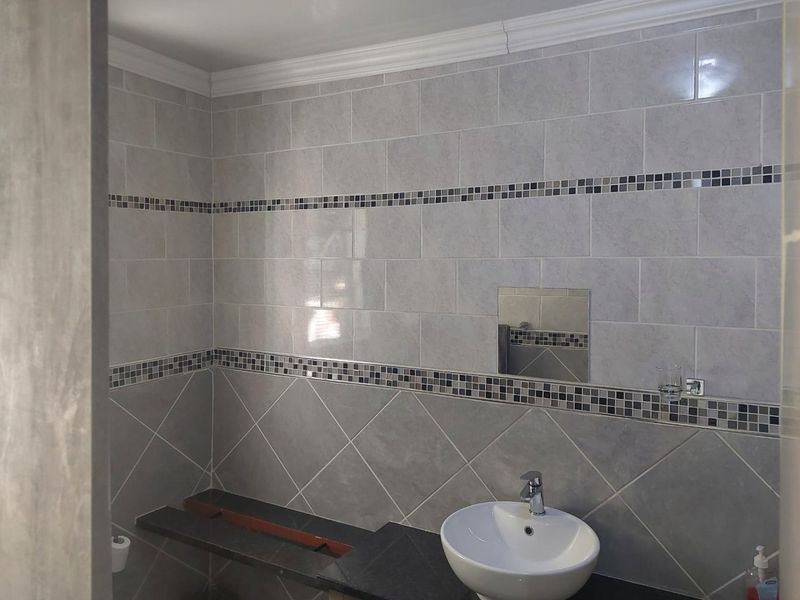 A bathroom featuring grey ceramic wall tiles with a mosaic border, a vessel sink on a stone vanity, and decorative crown molding.