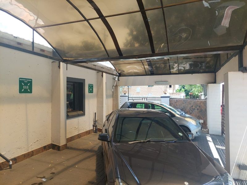 A covered parking area featuring a polycarbonate roof structure, paved flooring, and brick support pillars, providing sheltered vehicle storage.
