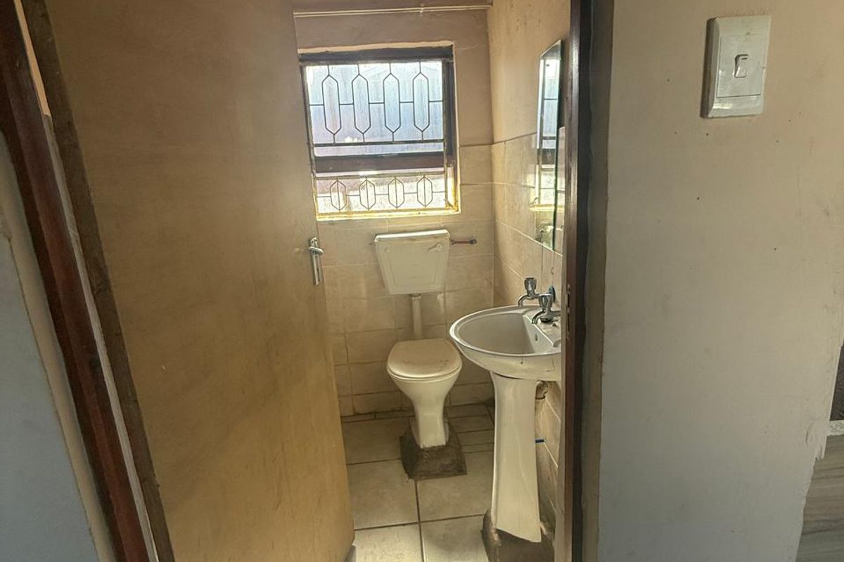 A compact, dated bathroom featuring a pedestal basin, toilet, tiled flooring, and a timber ceiling, requiring cosmetic updates.