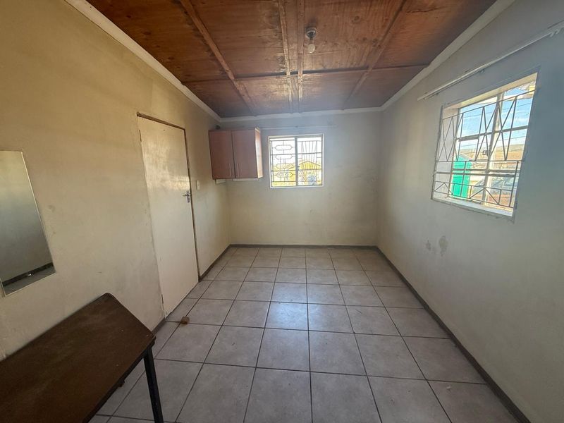 A simple, empty bedroom featuring tiled flooring, a wooden ceiling, and security bars on the windows, offering a blank canvas for renovation.