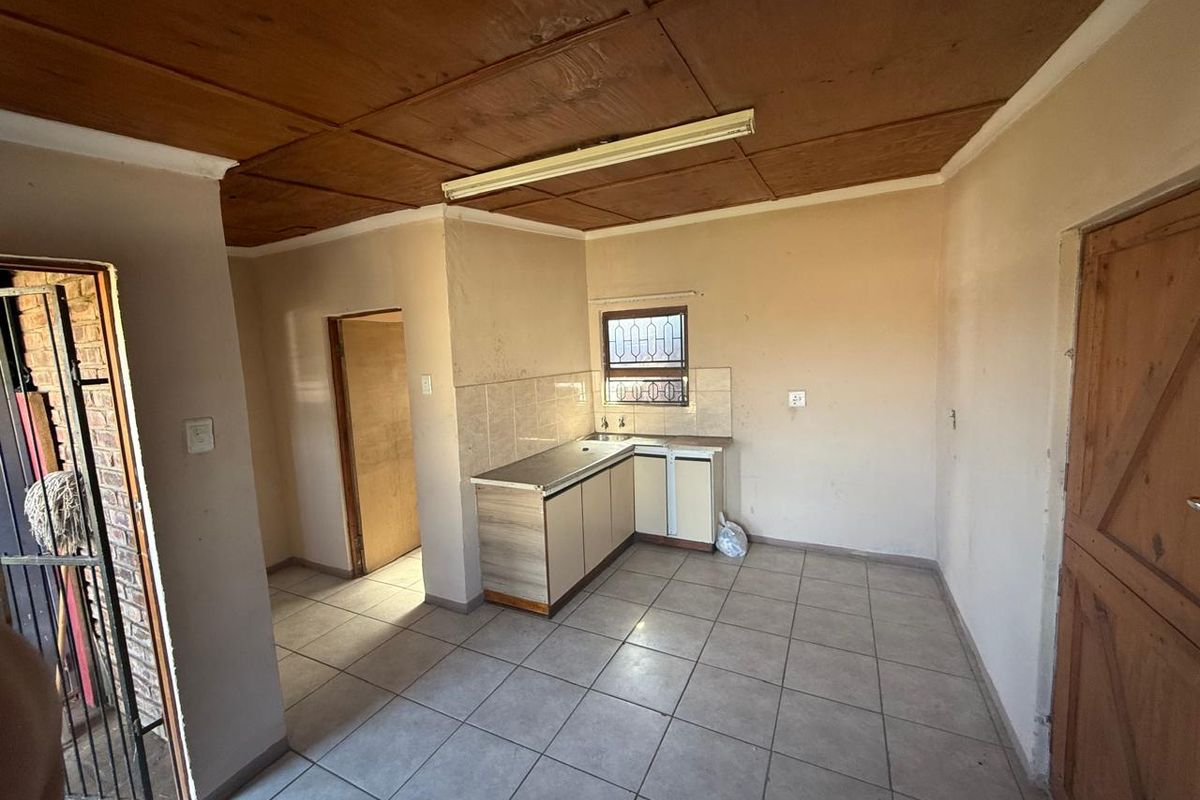 A compact, empty kitchen featuring tiled flooring, built-in cabinetry with a stainless steel sink, and a window with security bars.