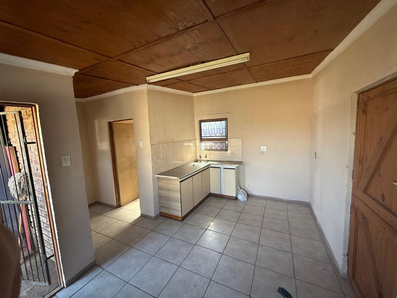 A compact, empty kitchen featuring tiled flooring, built-in cabinetry with a stainless steel sink, and a window with security bars.