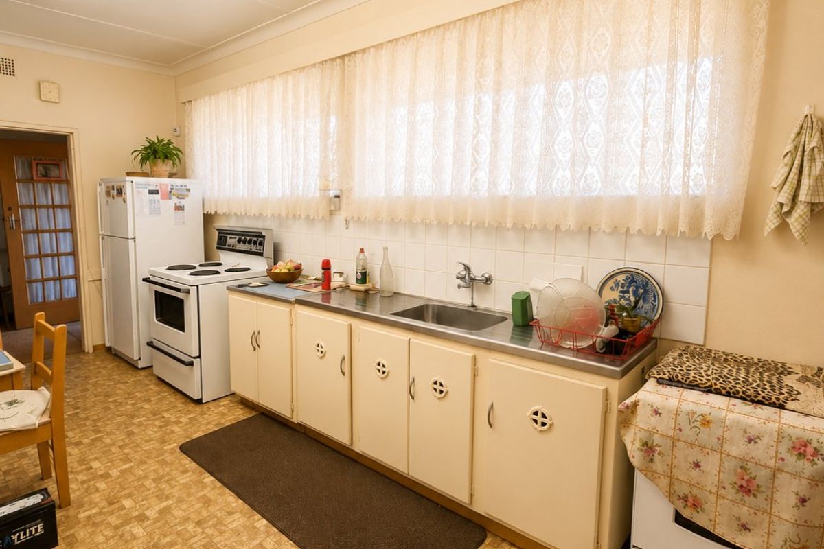 A functional, dated kitchen featuring original cabinetry, stainless steel sink, tiled splashback, and vinyl flooring, offering significant potential for modern renovation.