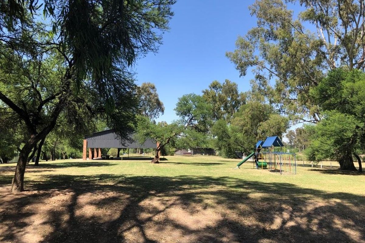 A spacious, well-maintained garden area featuring mature trees, a grass lawn, a playground structure, and an open-sided pavilion for outdoor recreation.