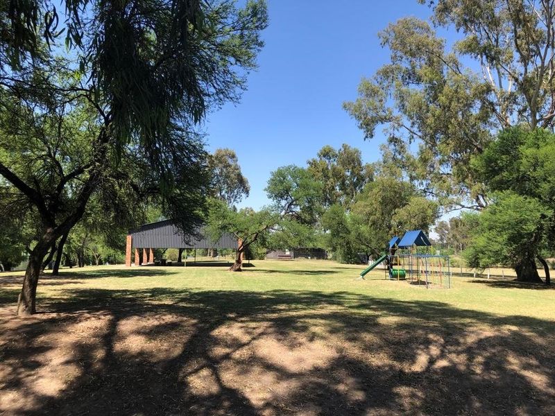 A spacious, well-maintained garden area featuring mature trees, a grass lawn, a playground structure, and an open-sided pavilion for outdoor recreation.