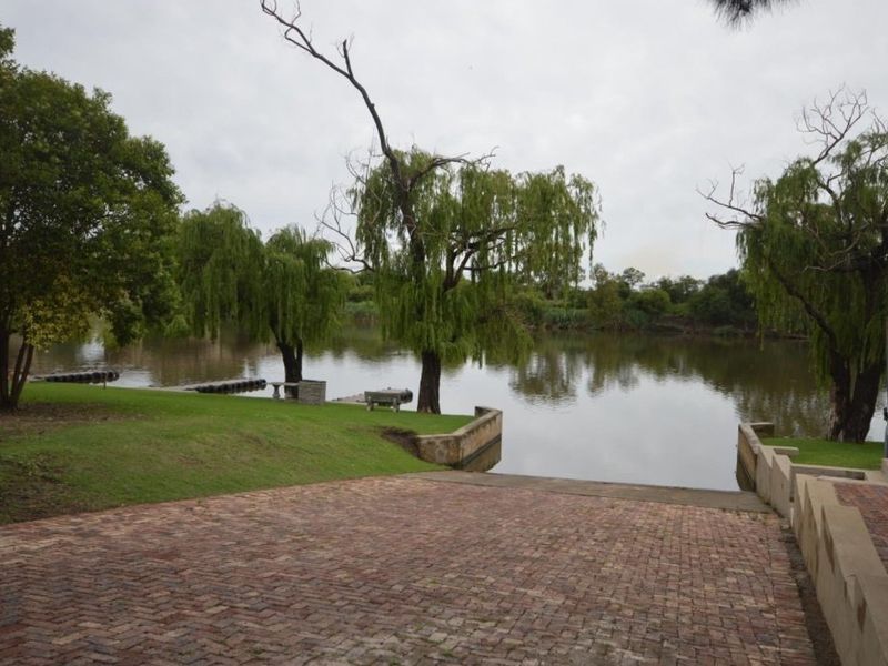 A scenic view of the riverfront featuring a brick-paved boat ramp, retaining walls, and mature willow trees along the water's edge.