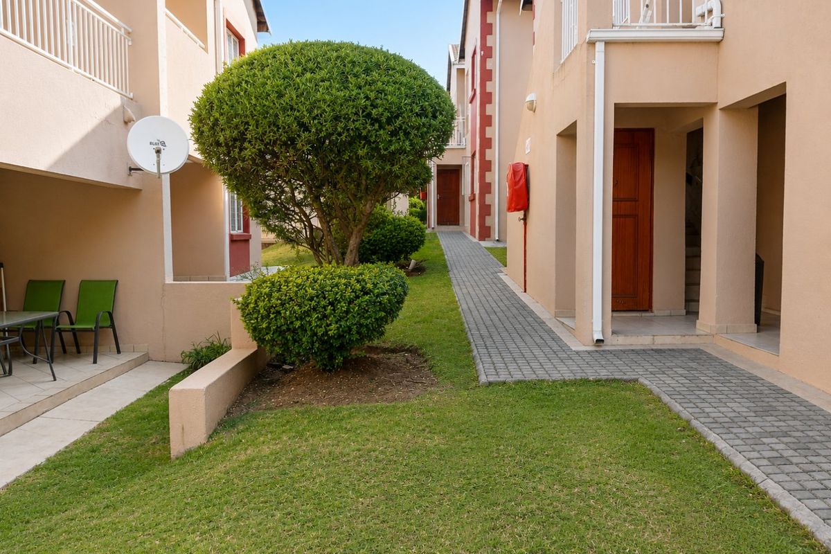 A view of the apartment complex exterior featuring a paved walkway, landscaped garden area, and access to ground floor units with private patio spaces.