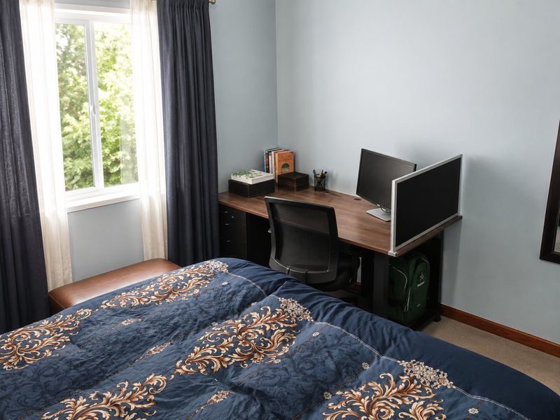 A bedroom featuring carpeted flooring, a window with curtains, and baseboard trim, currently utilized as a combined sleeping and workspace area.