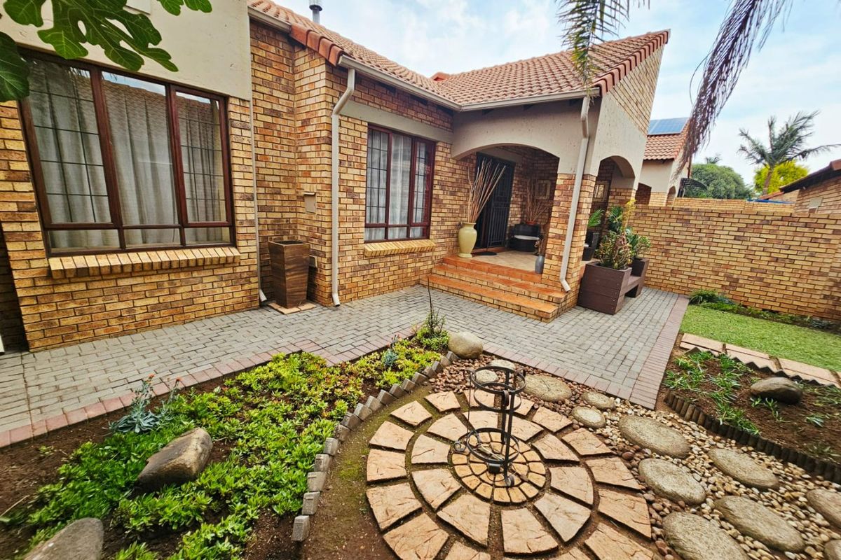 A well-maintained townhouse rear exterior featuring a face brick facade, covered entrance, paved patio area, and a landscaped garden with stone pathways.