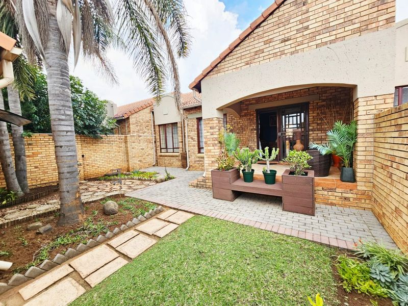 A well-maintained rear exterior view of a face brick townhouse featuring a covered patio, paved walkway, and landscaped garden area.