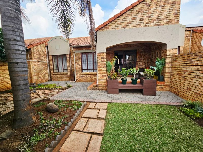 A well-maintained rear exterior view of a brick townhouse featuring a covered patio, paved walkway, and landscaped garden area.
