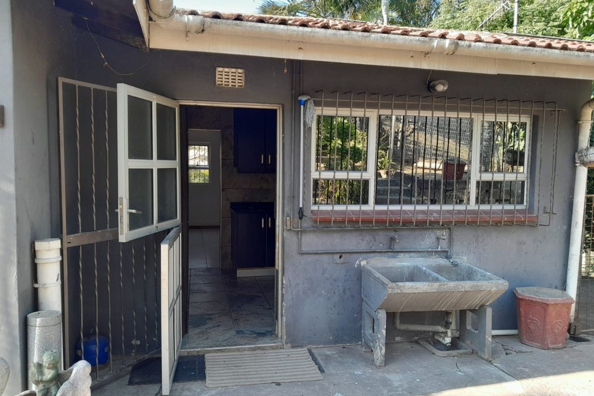 Rear exterior view showing a utility area with a concrete laundry trough, security bars on windows, and access to the interior kitchen space.