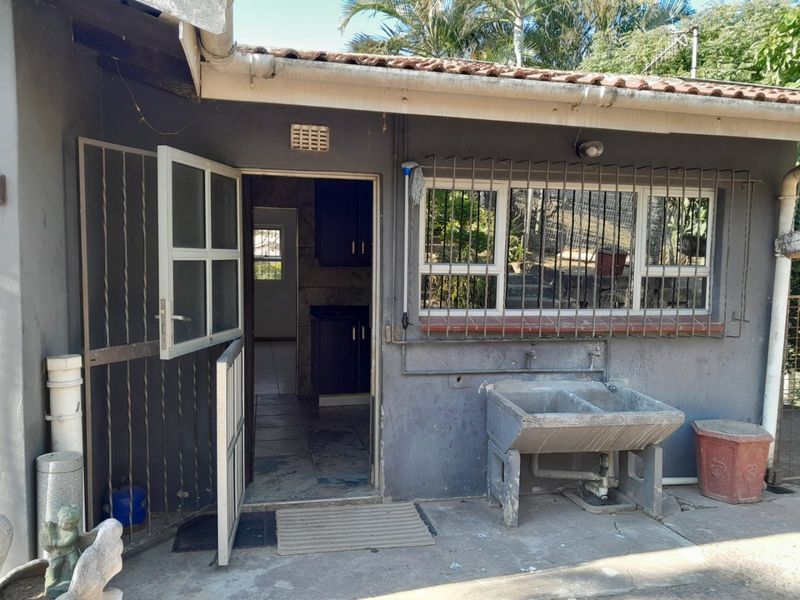 Rear exterior view showing a utility area with a concrete laundry trough, security bars on windows, and access to the interior kitchen space.
