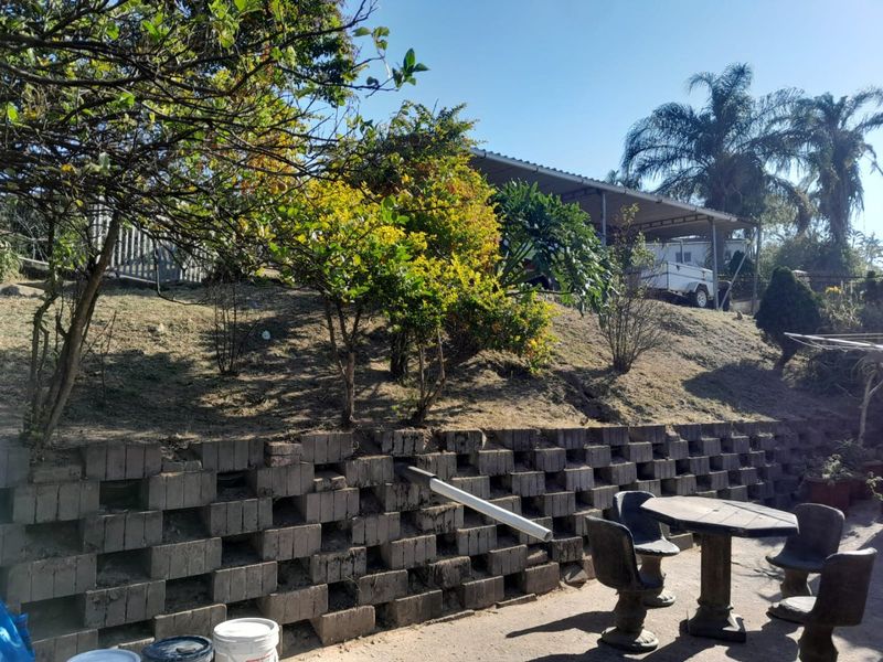 Rear exterior view featuring a tiered retaining wall, paved patio area, and an elevated carport structure with established garden landscaping.