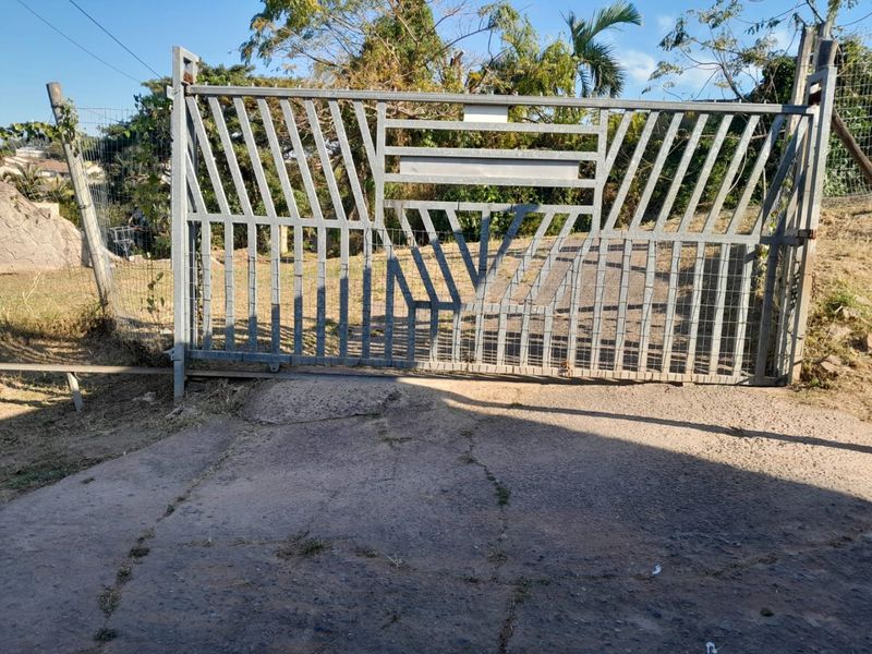 A metal security gate at the property entrance with an adjacent wire mesh fence and a weathered concrete driveway.