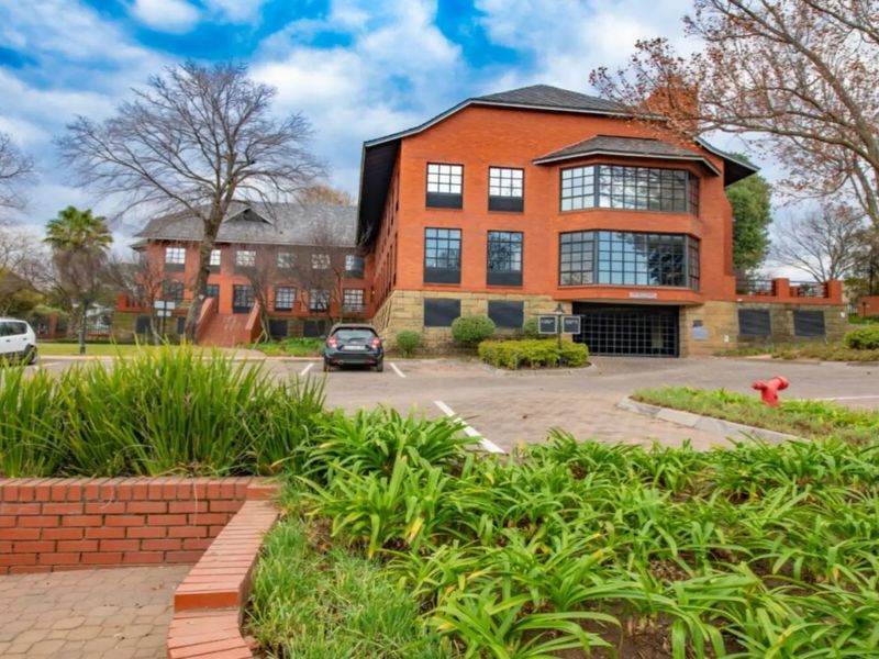 A professional exterior view of a multi-story commercial brick building featuring large multi-pane windows, a paved parking area, and established landscaped gardens.
