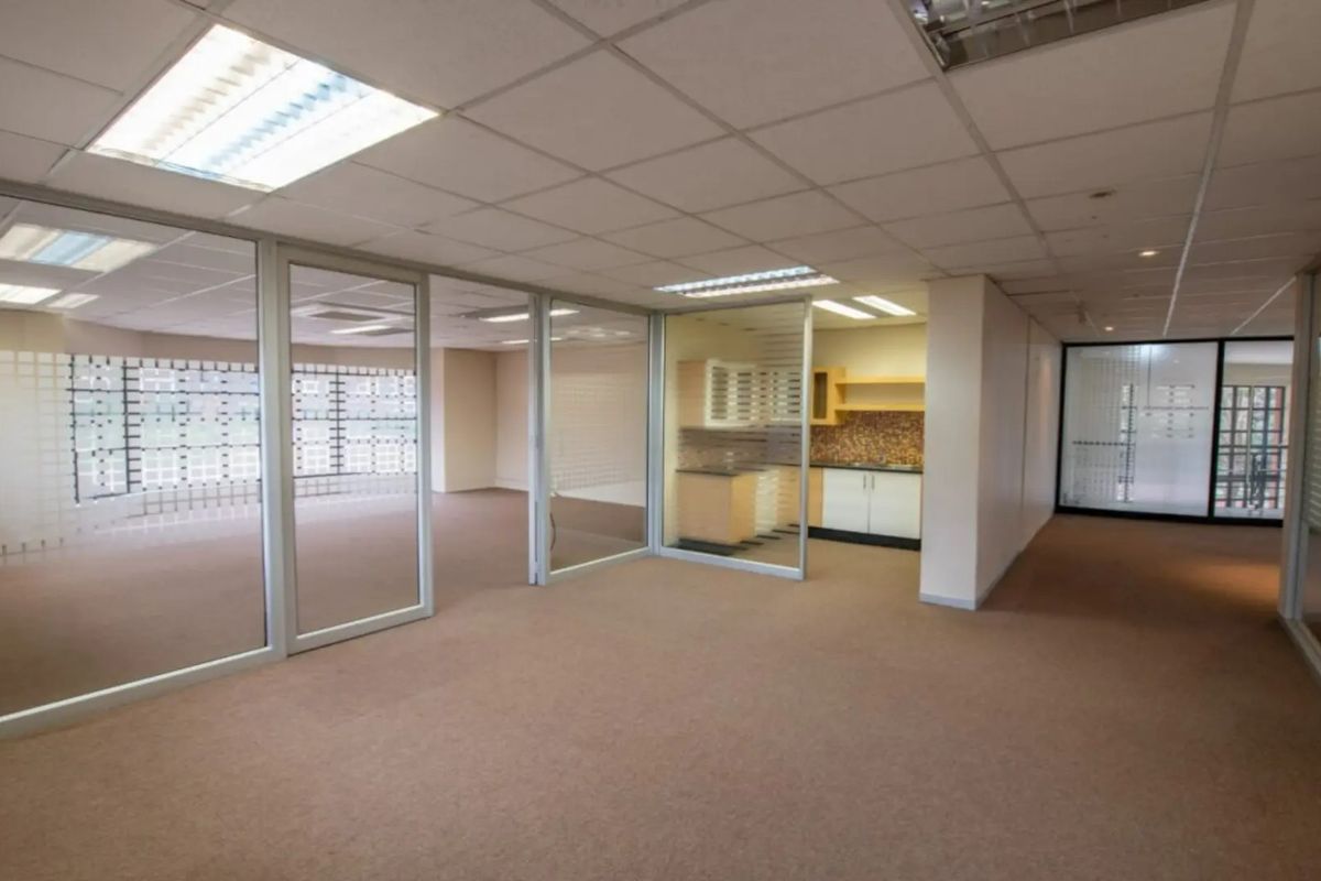Open-plan office space featuring glass partitions, a small kitchenette, suspended ceilings with recessed lighting, and neutral carpeting throughout.