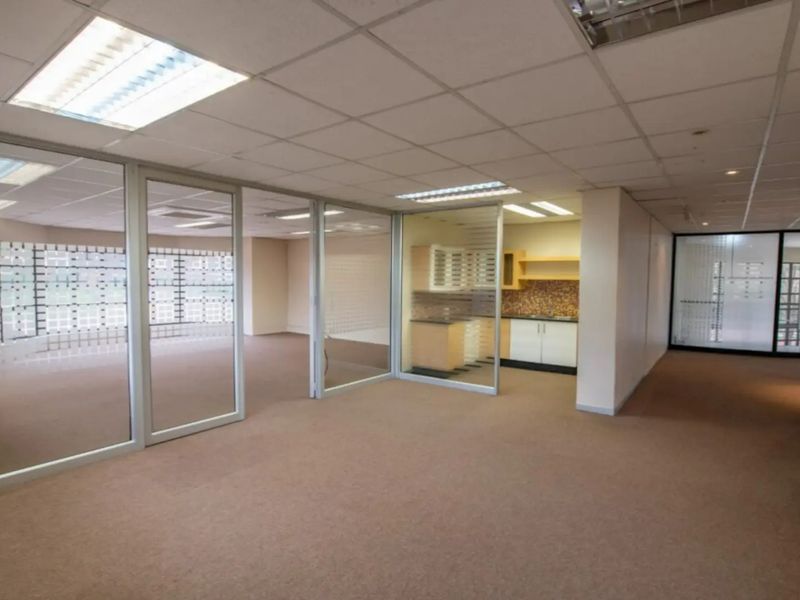 Open-plan office space featuring glass partitions, a small kitchenette, suspended ceilings with recessed lighting, and neutral carpeting throughout.