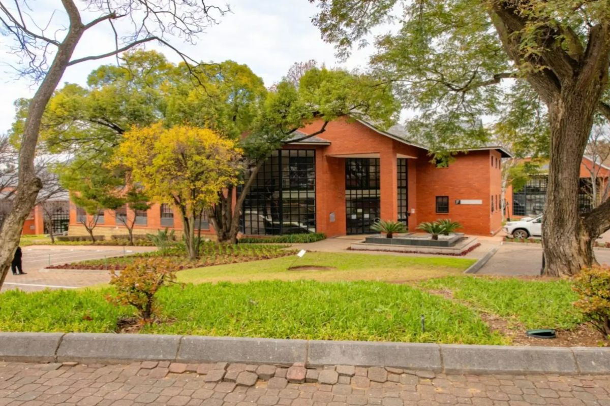 A professional exterior view of a commercial brick building featuring large glass windows, surrounded by mature trees and a well-maintained landscaped garden.