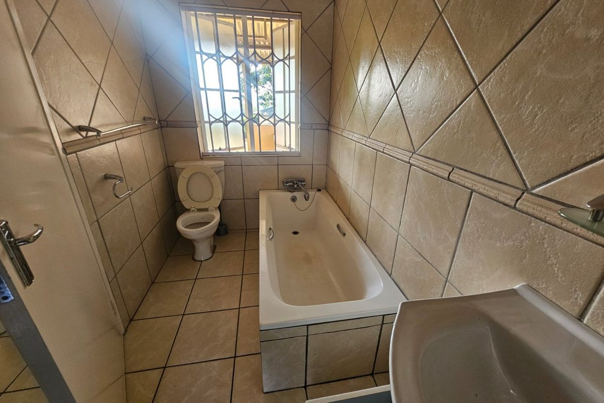 A dated bathroom featuring a built-in bathtub, toilet, and pedestal basin with full-height wall tiling and security bars on the window.