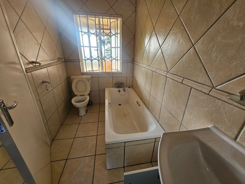 A dated bathroom featuring a built-in bathtub, toilet, and pedestal basin with full-height wall tiling and security bars on the window.